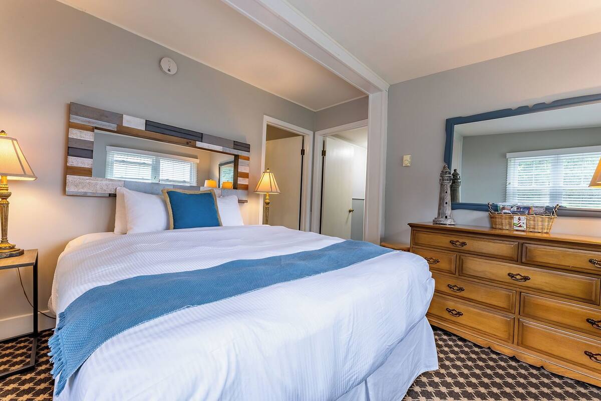 A hotel room with a large bed, a wooden dresser, a mirror, and a window with blinds.