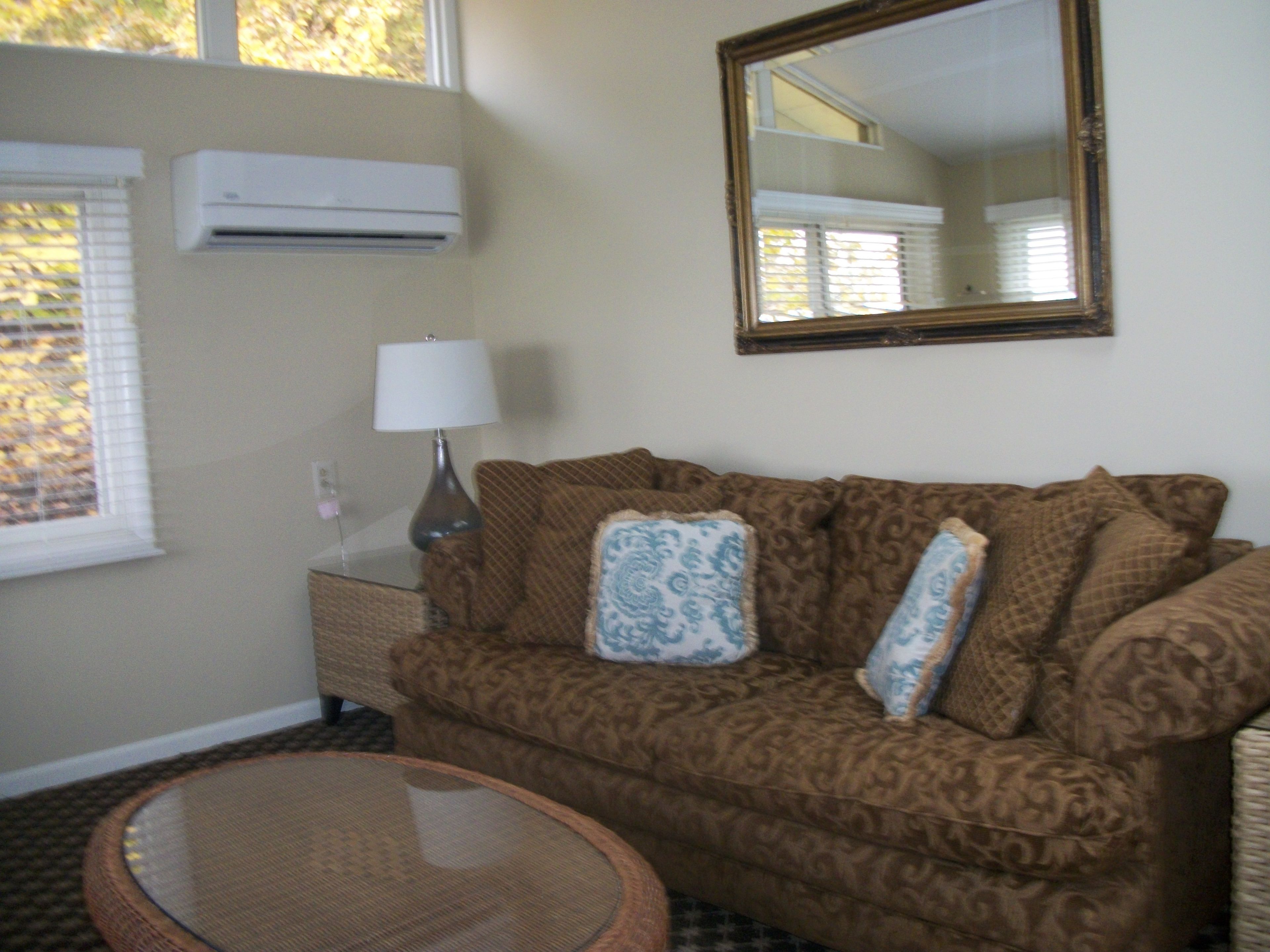 A living room with a patterned sofa, a round glass coffee table, a lamp, and a large mirror on the wall.