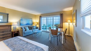 A hotel room with a bed, a sofa, a dining table with chairs, a large window with curtains, and a painting on the wall.