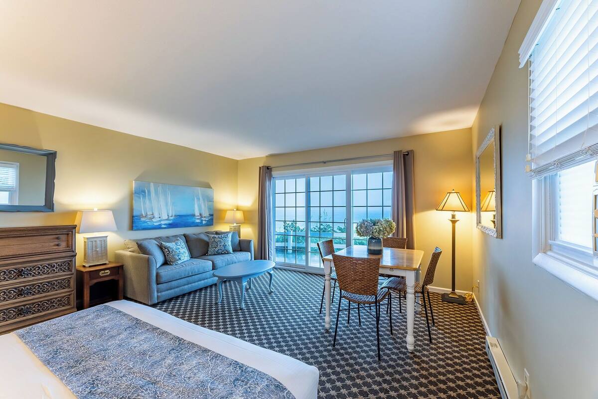 A hotel room with a bed, a sofa, a dining table with chairs, a large window with curtains, and a painting on the wall.