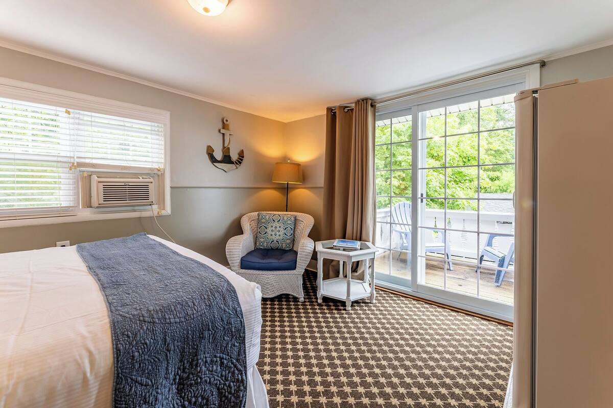 A hotel room with a bed, a chair, a small table, a lamp, and a large window with a view of the outdoors.