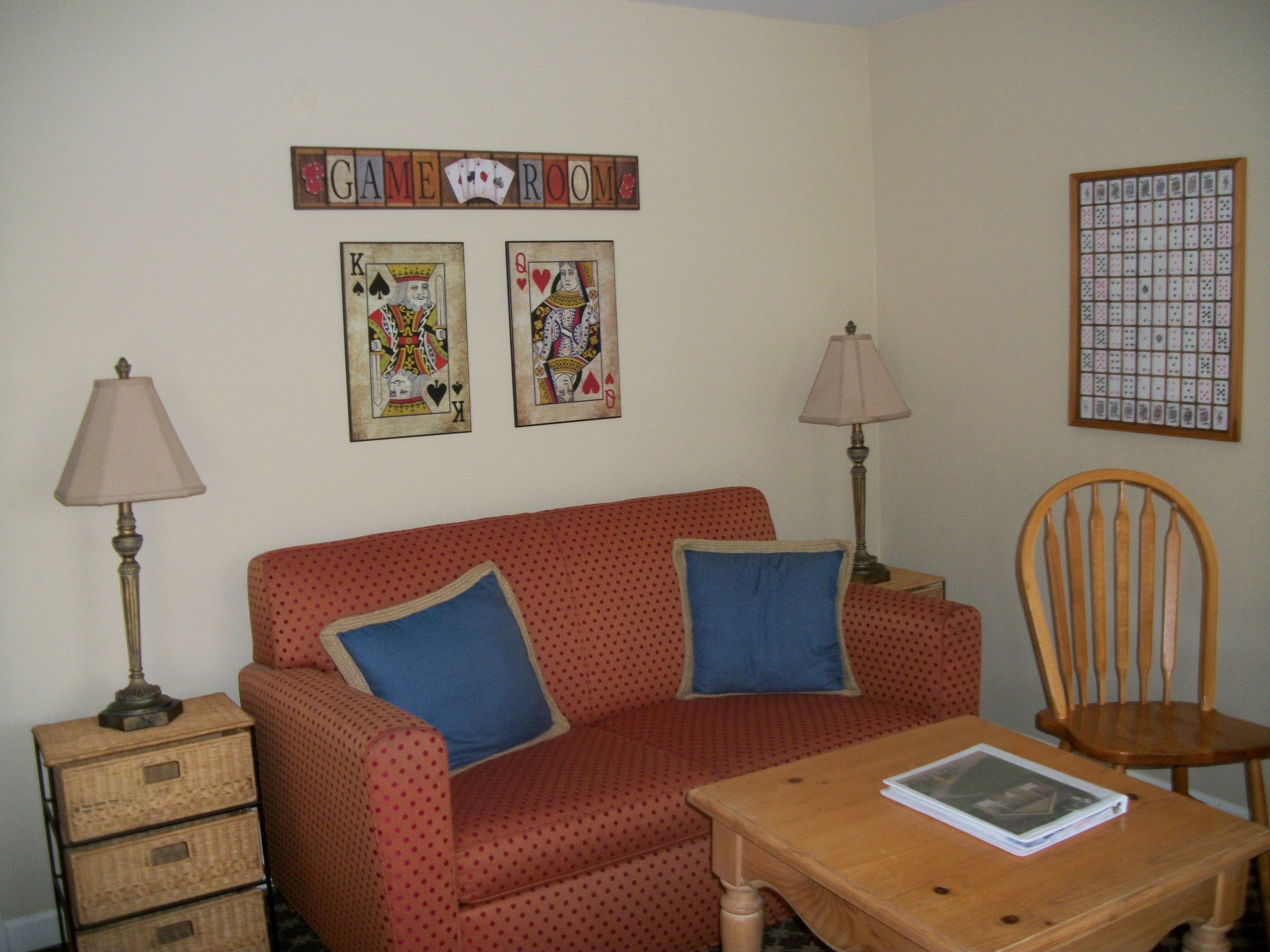 A living room with a red sofa, two blue cushions, a wooden table with a book, two lamps, and a wall-mounted card game board.