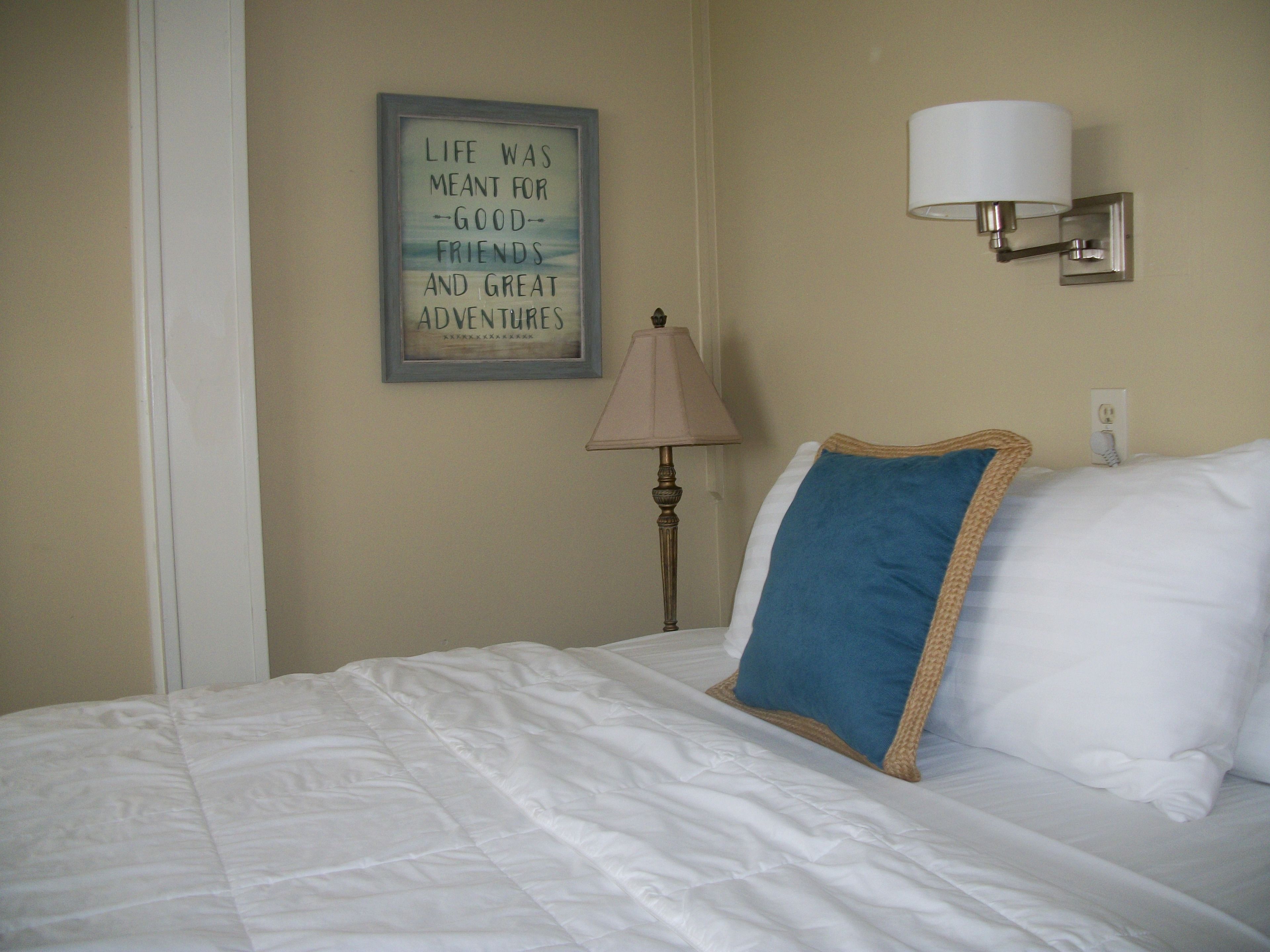 Una cama bien hecha con ropa blanca y una almohada azul, una lámpara de pared y una cita inspiradora enmarcada.