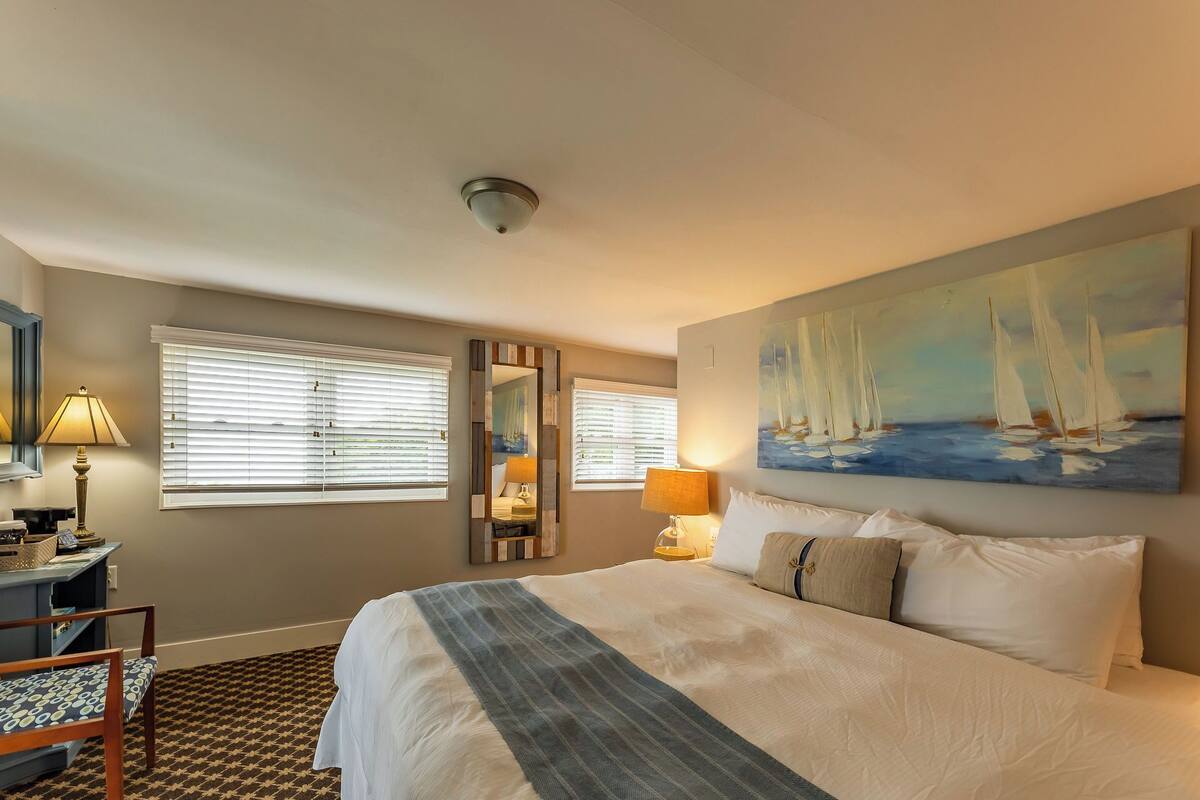 A hotel room with a large bed, two bedside lamps, a painting of sailboats, a mirror, and a small table with a lamp.