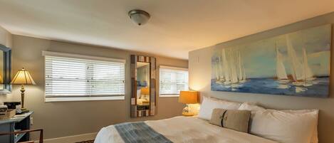 A hotel room with a large bed, two bedside lamps, a painting of sailboats, a mirror, and a small table with a lamp.