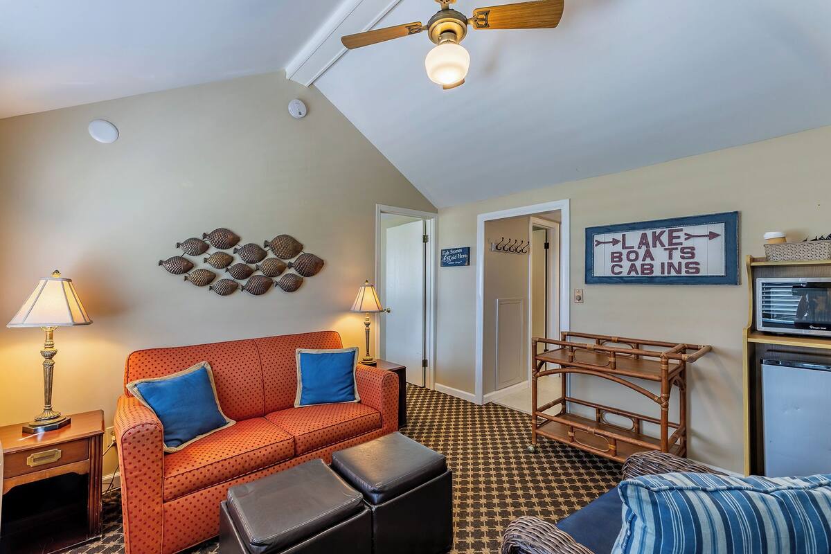 A cozy living room with a red sofa, a wooden chair, and a small table. There's a sign on the wall indicating 'Lake Boats Cabins'.