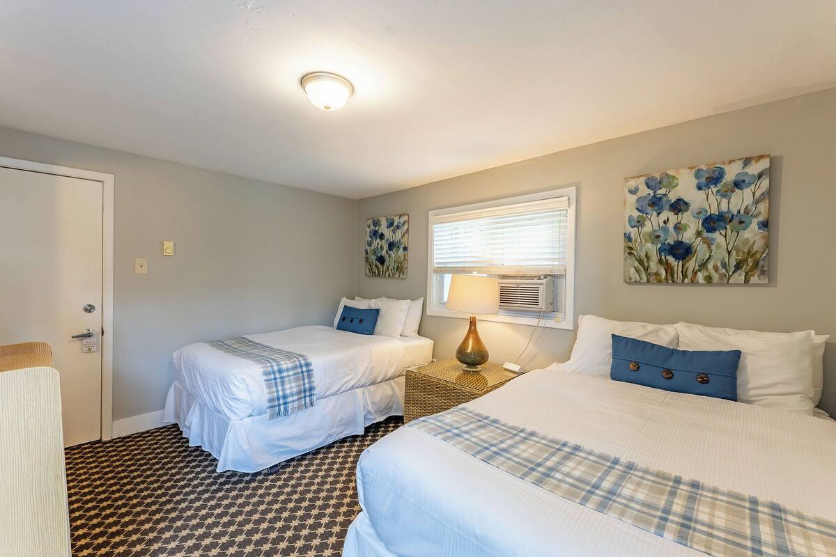 A hotel room with two beds, a window with blinds, a wall-mounted air conditioner, and a painting of flowers.
