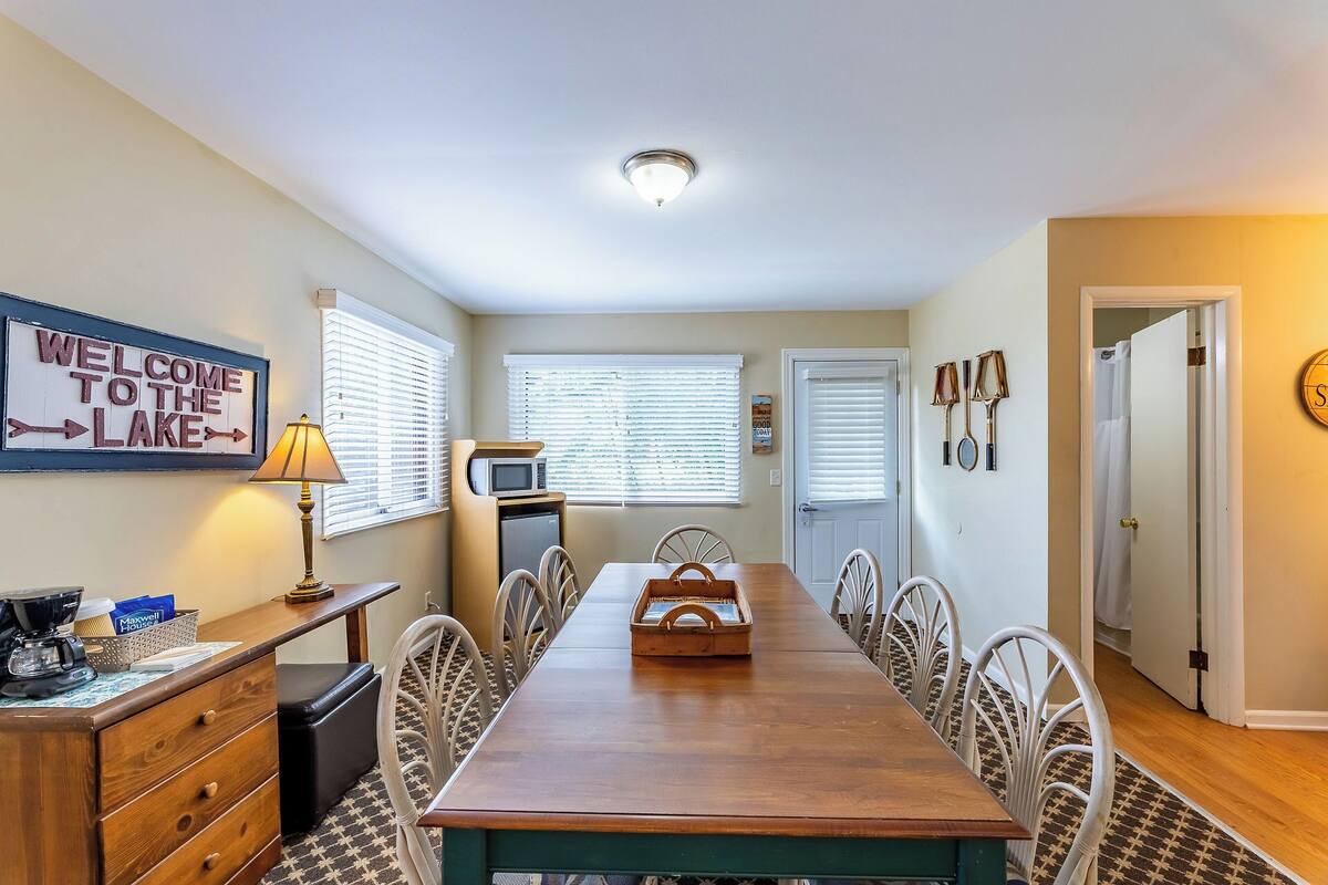 A dining area with a long wooden table, white chairs, a microwave, and a sign that reads "WELCOME TO THE LAKE".