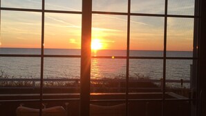 A sunset view through a window with a grid pattern, overlooking a calm sea and a wooden deck with chairs.