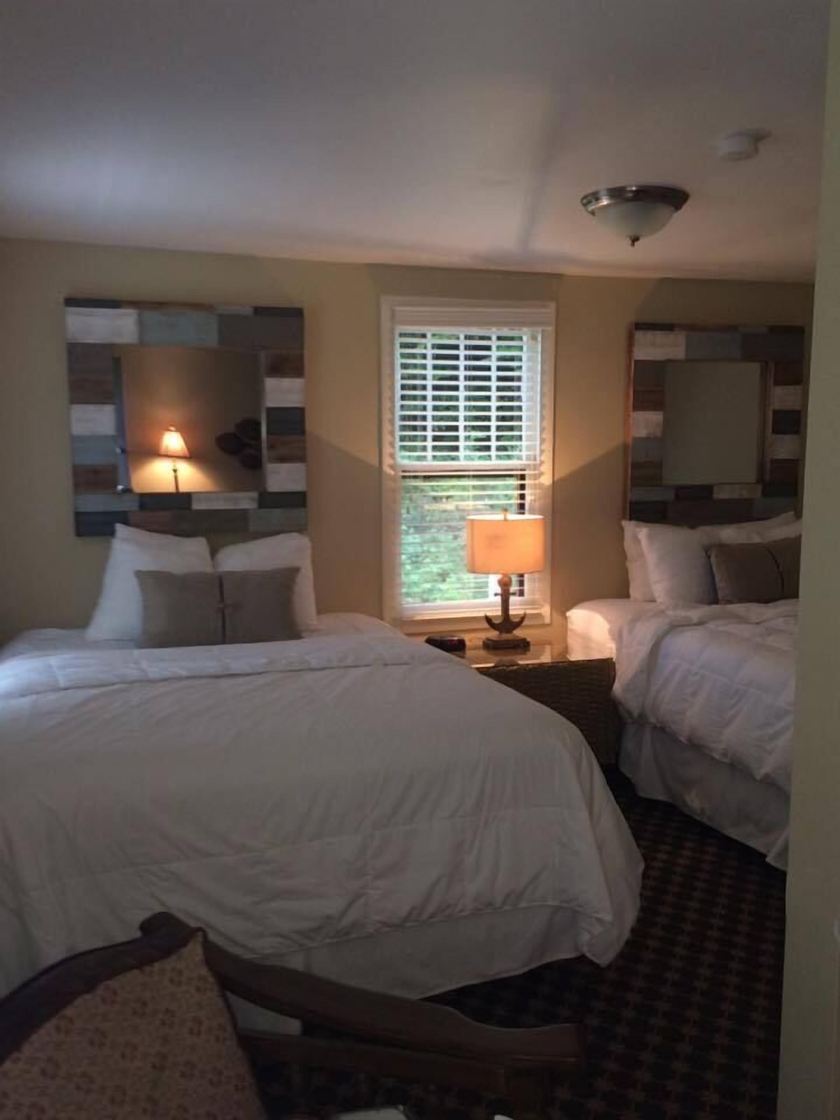 A hotel room with two beds, a window with blinds, a lamp on a nightstand, and a ceiling light.