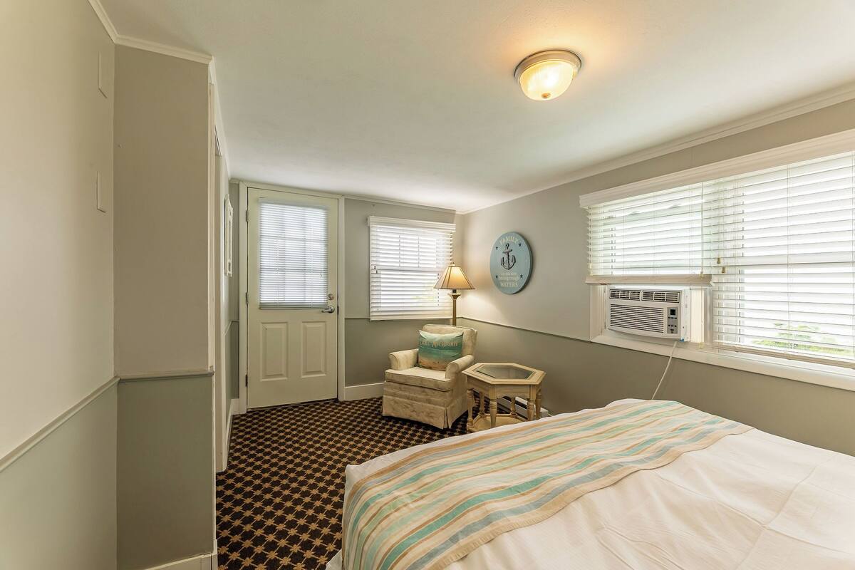 A bedroom with a bed, a chair, a lamp, a wall clock, and an air conditioning unit.