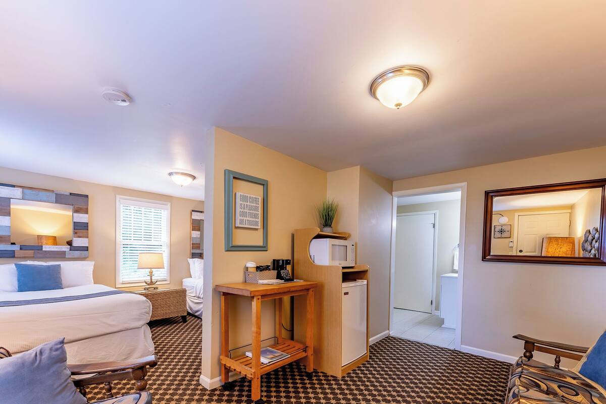 A hotel room with a bed, a desk, a refrigerator, a mirror, and a door leading to another room.