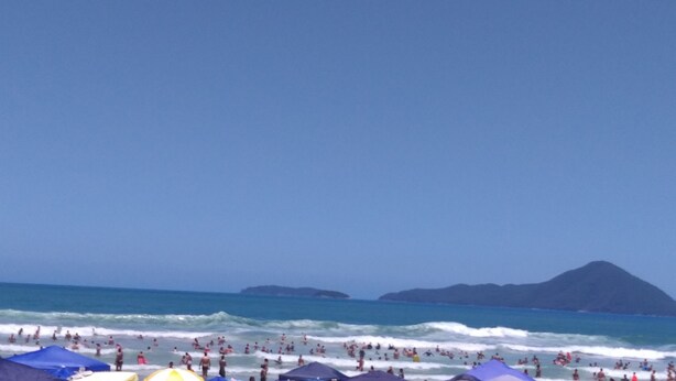 Una playa con gente nadando y tomando el sol, sombrillas y una montaña al fondo.