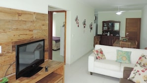 Una sala de estar con un televisor de pantalla plana sobre un soporte de madera, un sofá blanco con cojines florales, un ventilador de techo y una zona de cocina al fondo.