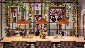 Un espace repas comprenant une longue table, six chaises et trois lampes suspendues. Les murs sont décorés de compositions florales et de plantes vertes.