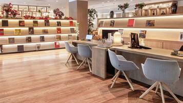 En modern hotellobby med en receptionsdisk i trä, grå stolar och en utställning med böcker och dekorativa föremål.