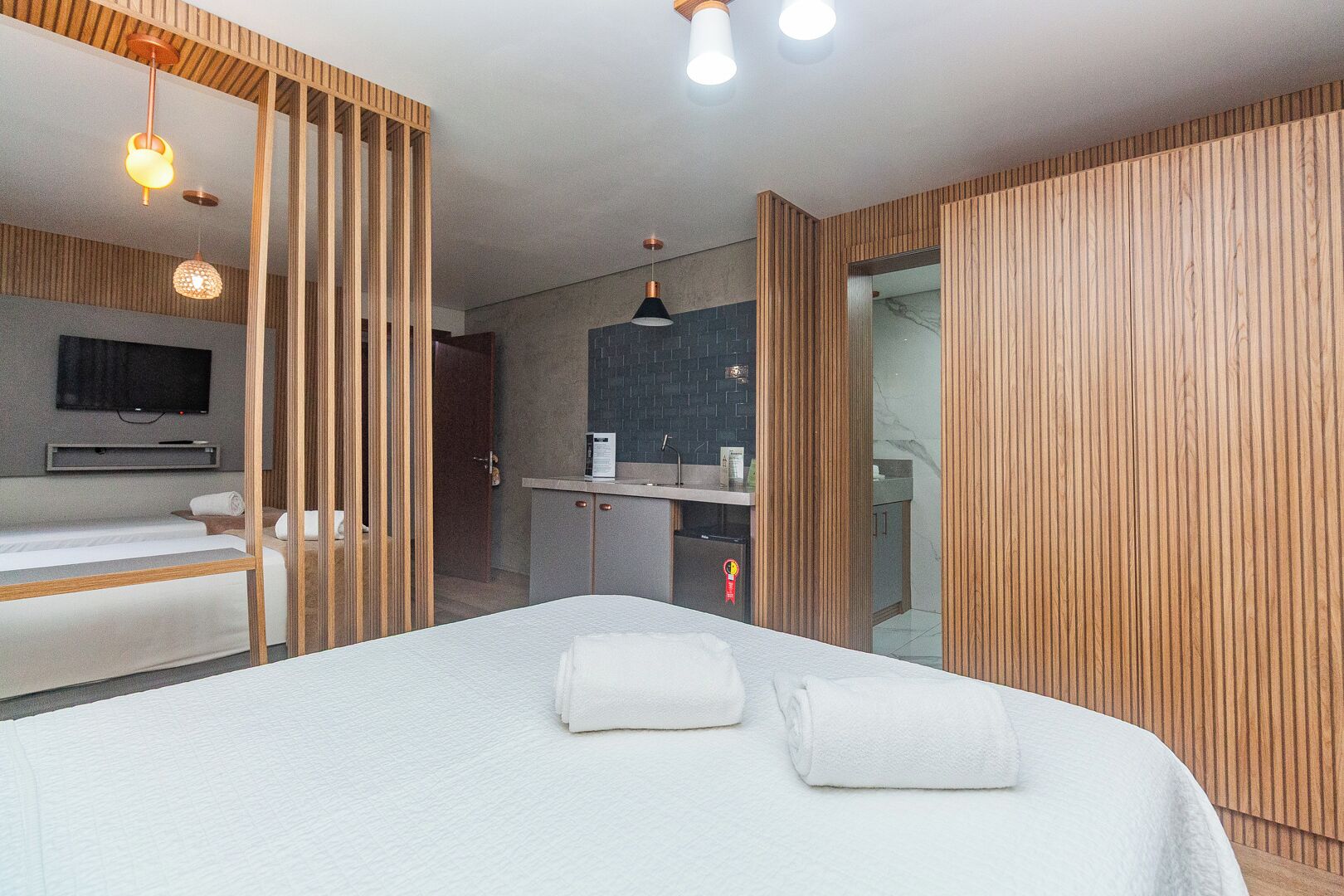 A modern hotel room with a large bed, a TV mounted on the wall, a small kitchenette, and wooden paneling.