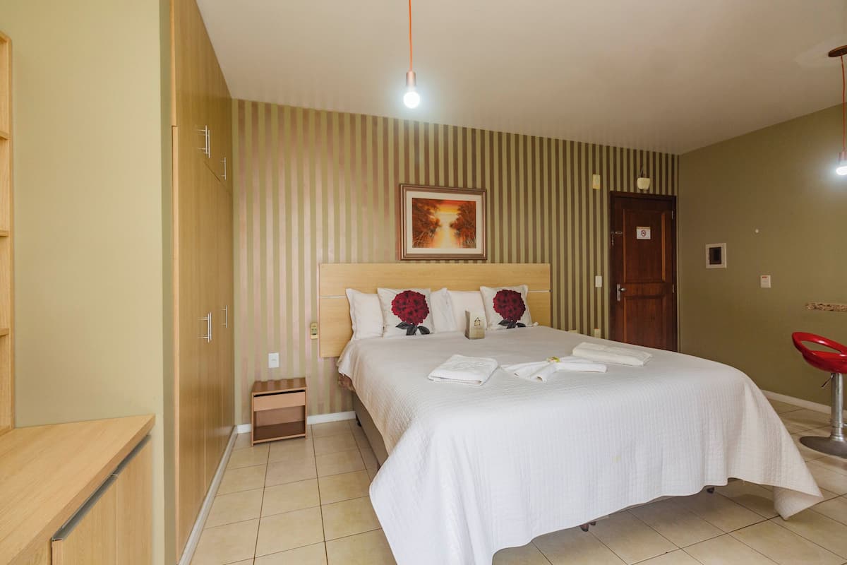 A hotel room with a bed, a nightstand, a wooden wardrobe, a red chair, and a painting on the wall.