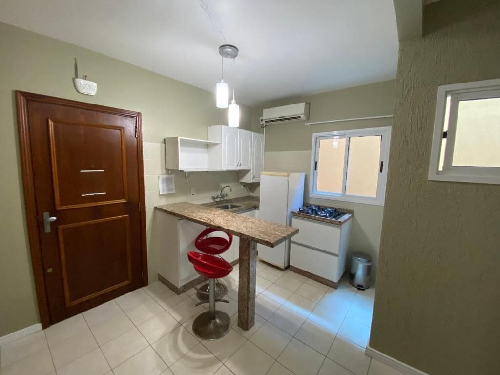 A compact kitchen with a wooden door, white cabinets, a small island with red bar stools, a window with blinds, and a white air conditioning unit.