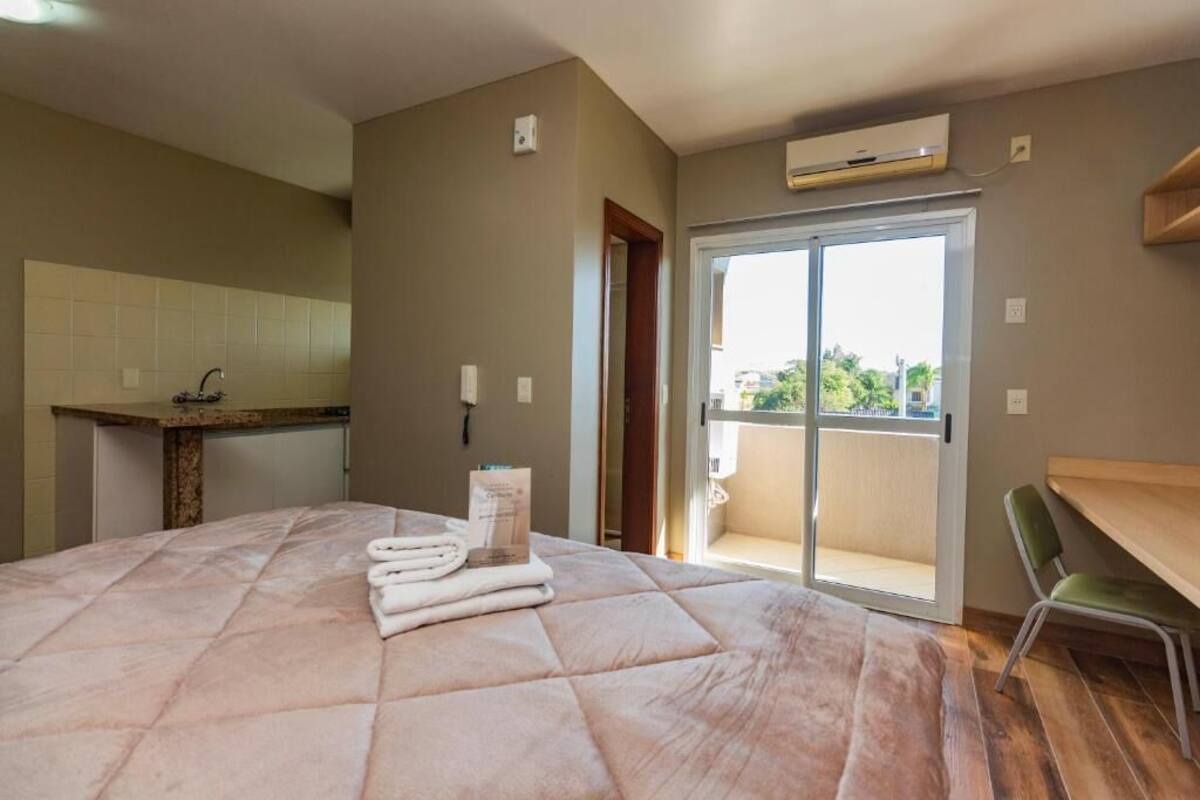 A hotel room with a bed, a small kitchenette, a balcony with a view, and a chair.