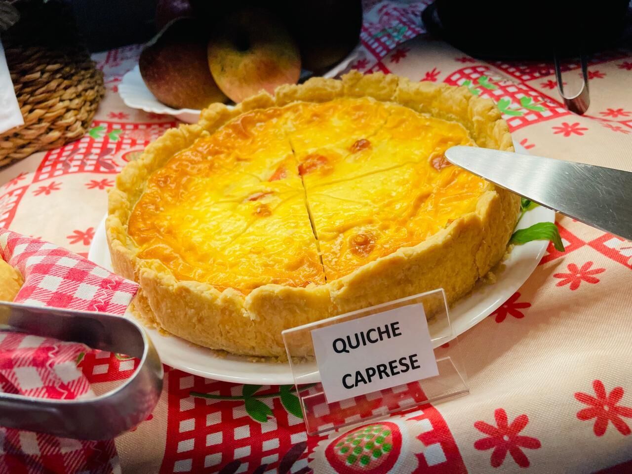 Una quiche sobre un plato blanco con una etiqueta, colocada sobre una mesa con mantel a cuadros rojo y blanco.