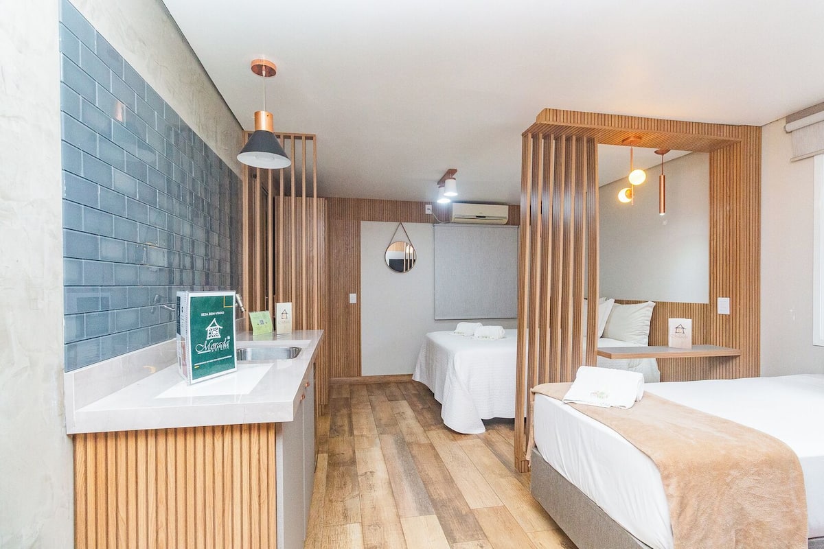 A hotel room with a wooden floor, two beds, a bathroom with a mirror, and a sink with a green sign.