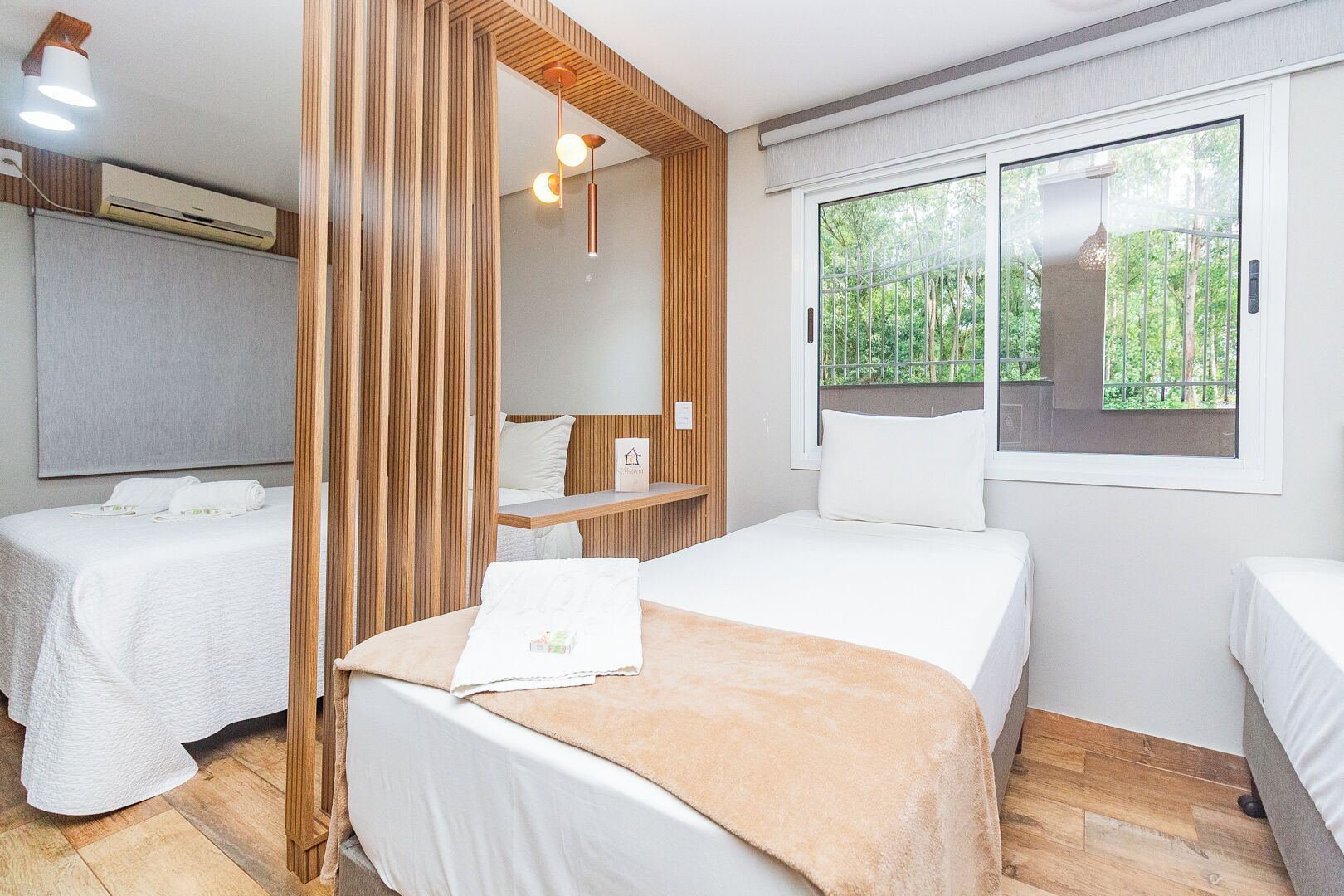 A hotel room with two beds, a wooden partition, a window with a view of greenery, and a hanging light fixture.