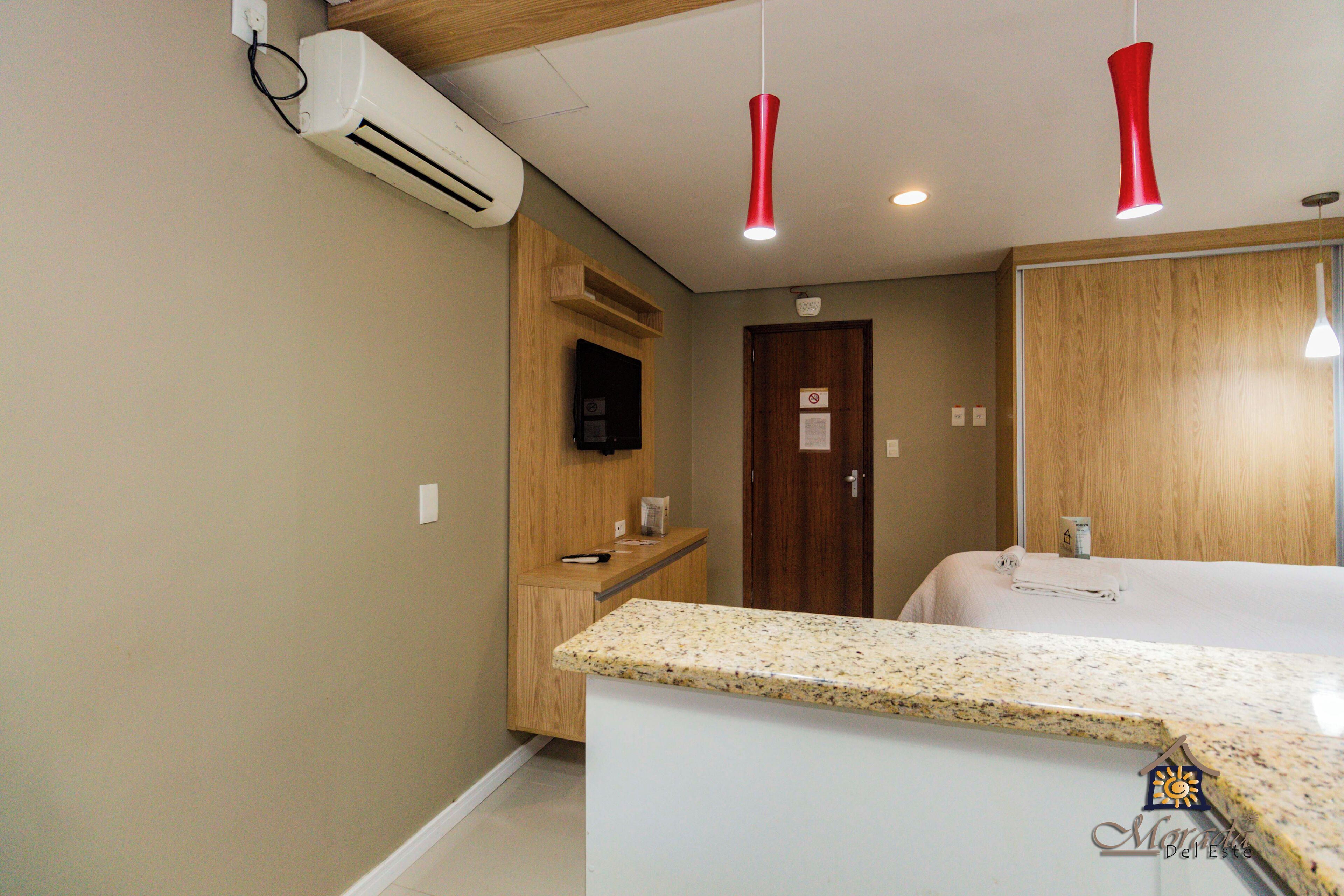 Habitación de hotel con kitchenette, un mueble de madera, un televisor, una puerta con un letrero y una cama con una toalla.