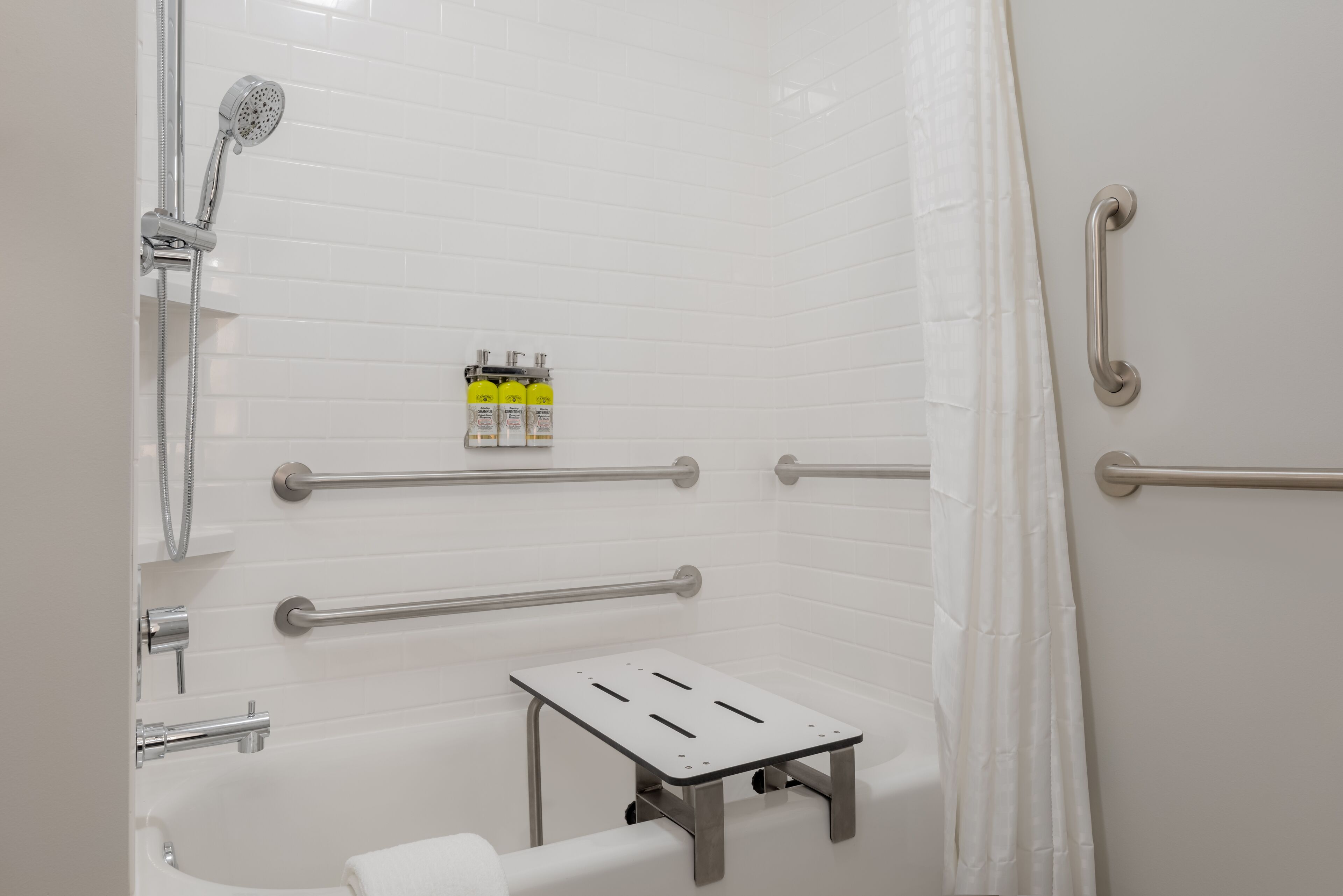 A modern bathroom with white tiles, a shower, grab bars, and a small table.