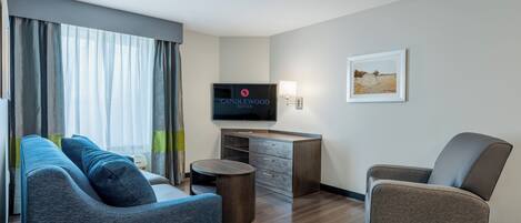 A modern hotel room with a sofa, a chair, a desk, a television, and a framed picture on the wall.