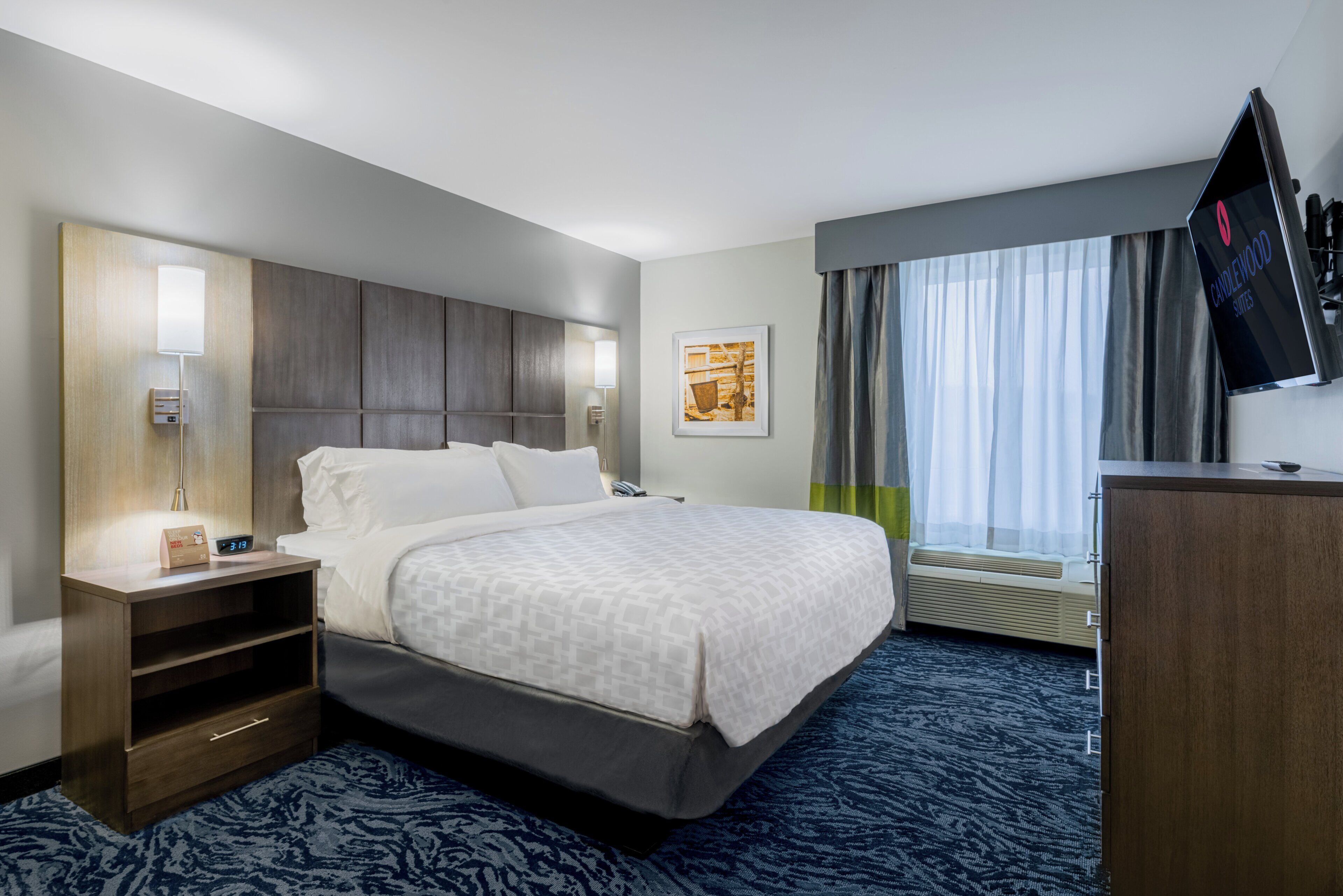 A hotel room with a bed, bedside table, lamp, artwork, and a TV on the wall.