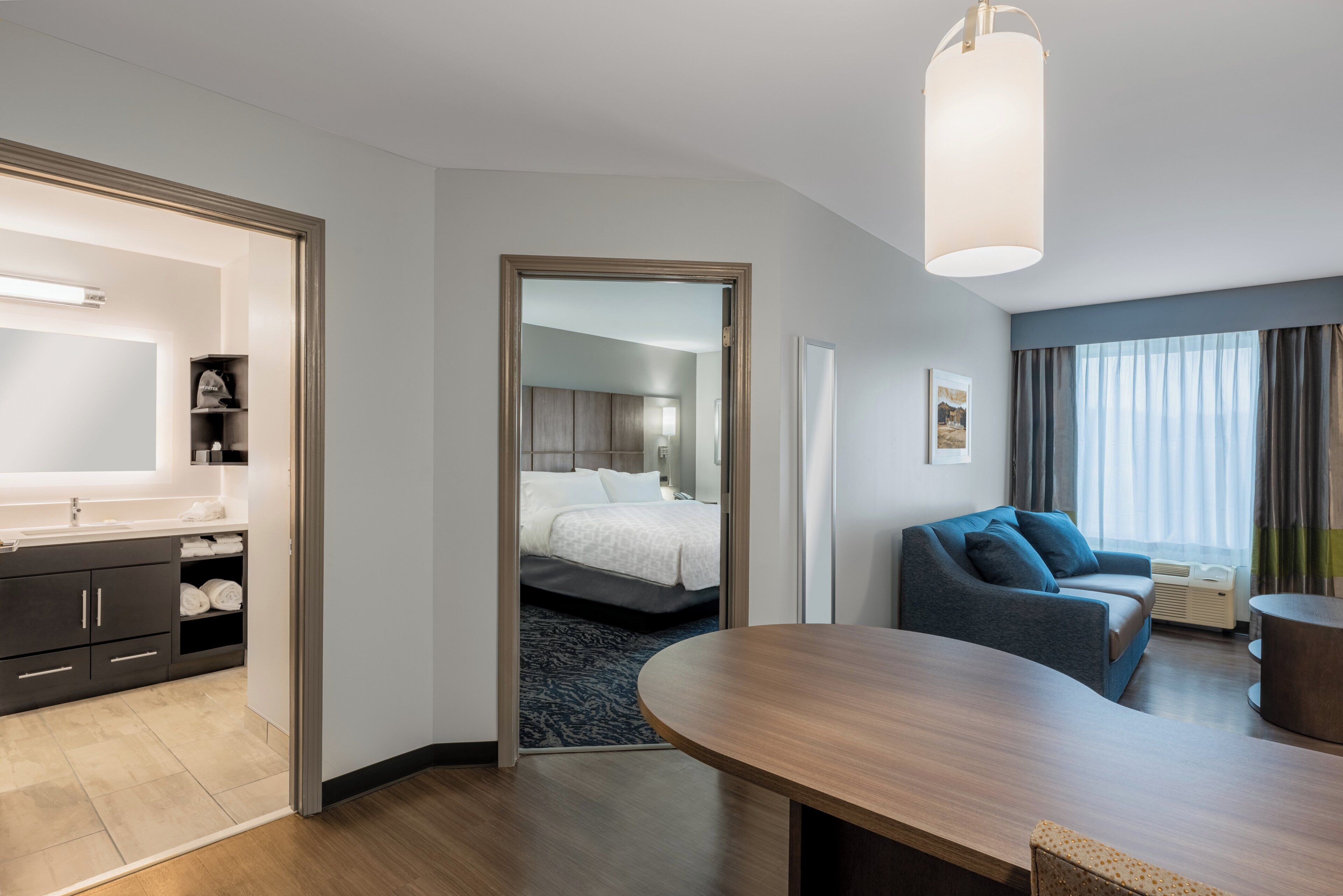 A modern hotel room with a wooden dining table, a sofa, a bed, and a bathroom with a mirror and towel rack.