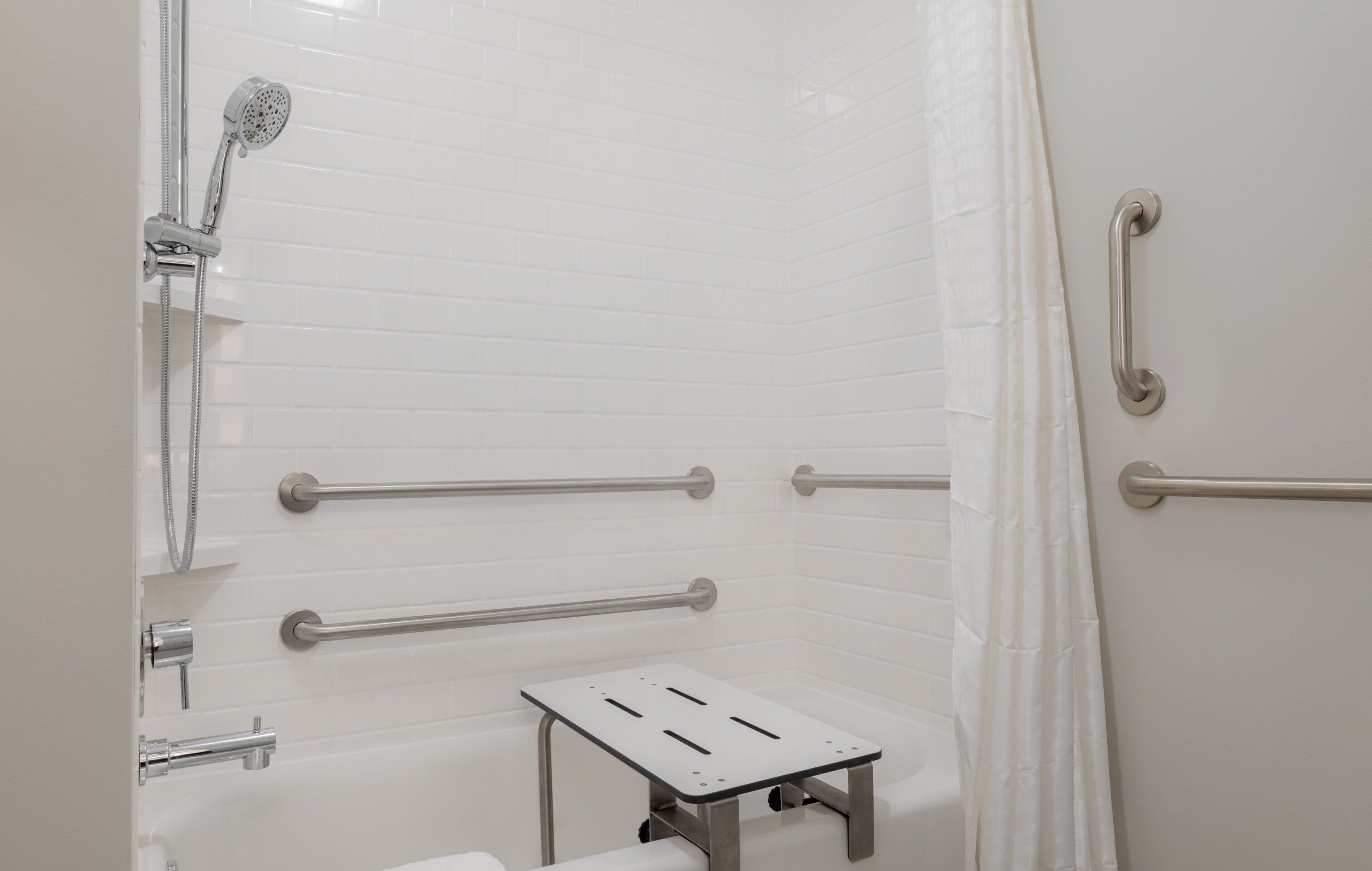 A modern bathroom with white tiled walls, a shower with a handheld showerhead, grab bars, and a small step stool.