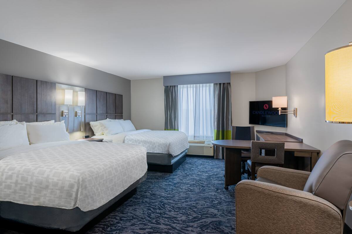 A hotel room with two beds, a desk, a chair, a TV, and a large window with curtains.