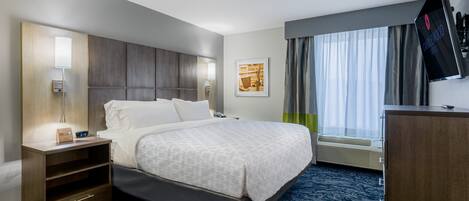 Habitación de hotel con cama, mesita de noche con lámpara, televisor de pantalla plana, ventana con cortinas y una obra de arte colgada en la pared.