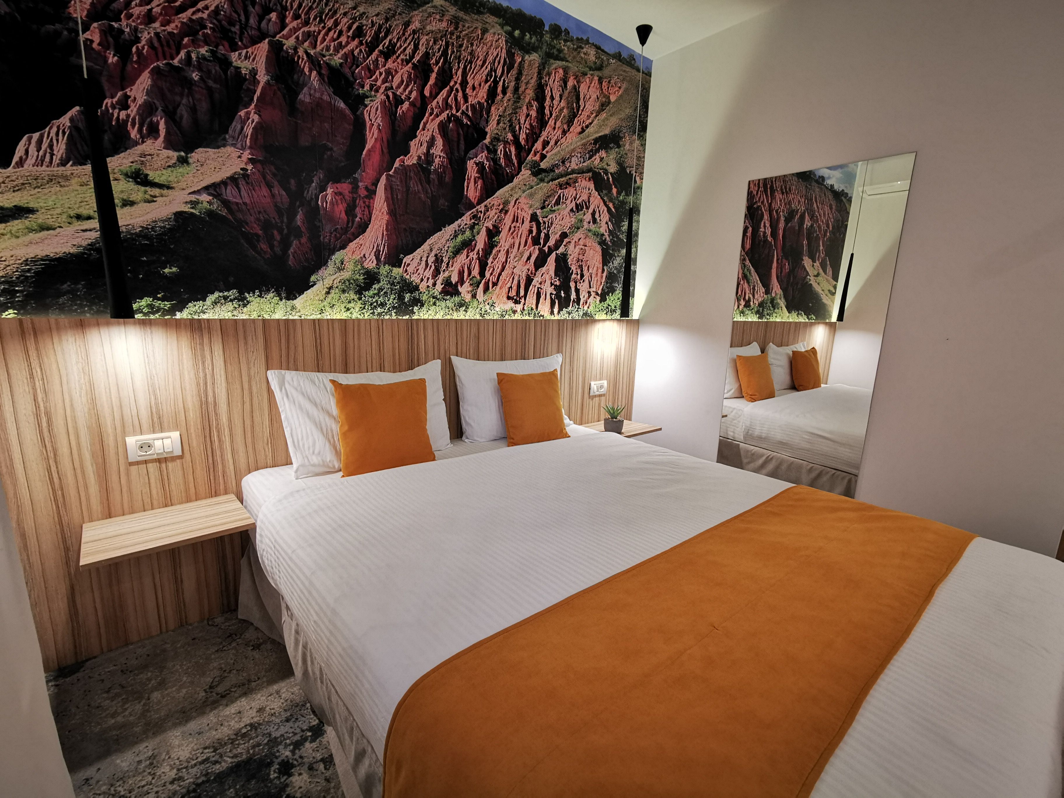 Habitación de hotel con una cama grande, dos mesitas de noche, un espejo y un mural en la pared que muestra un paisaje de rocas rojas.