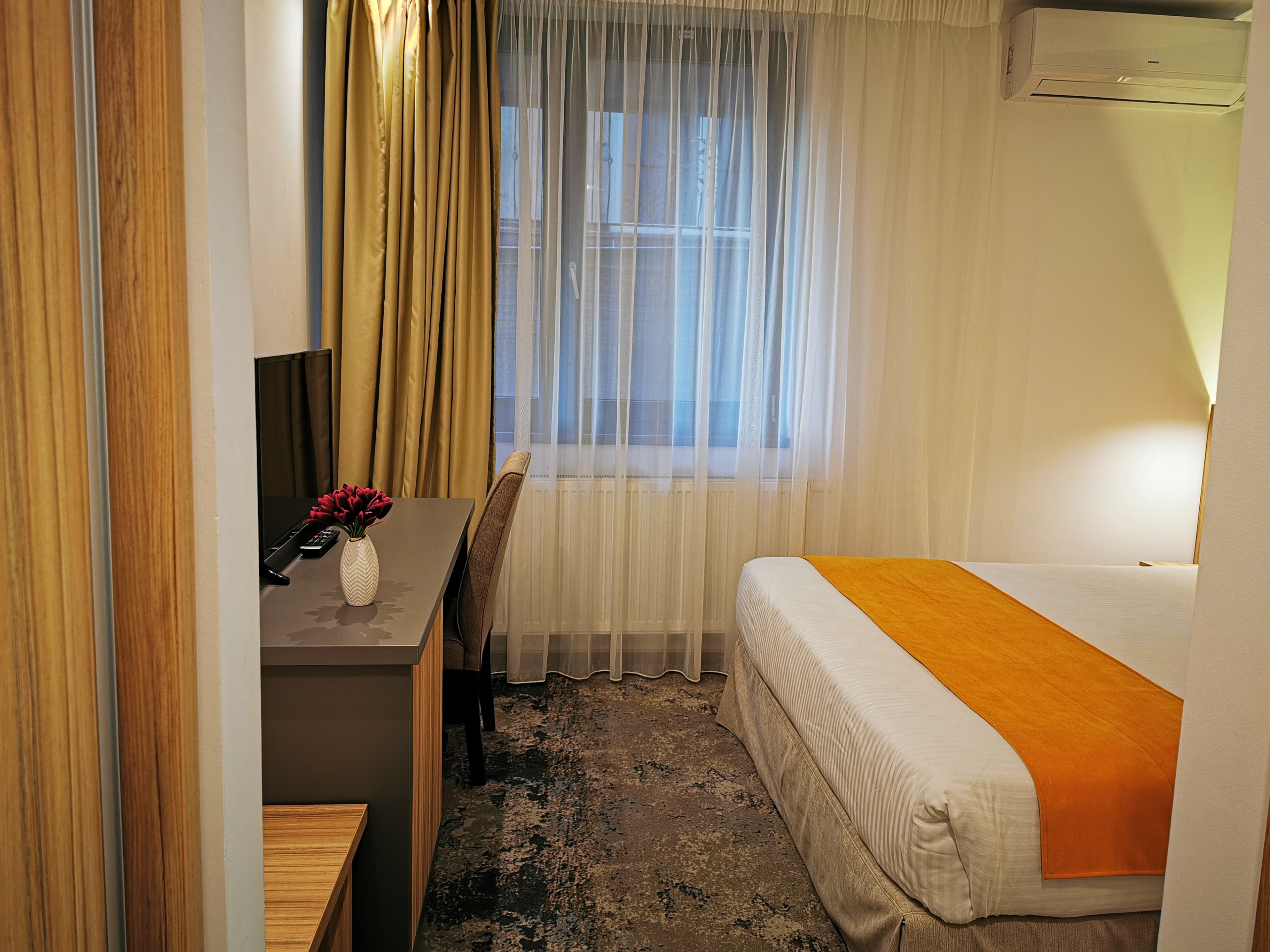 A hotel room with a bed, a desk, a chair, a small table with a vase and remote controls, and a window with sheer curtains.