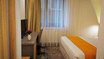 A hotel room with a bed, a desk, a chair, a small table with a vase and remote controls, and a window with sheer curtains.