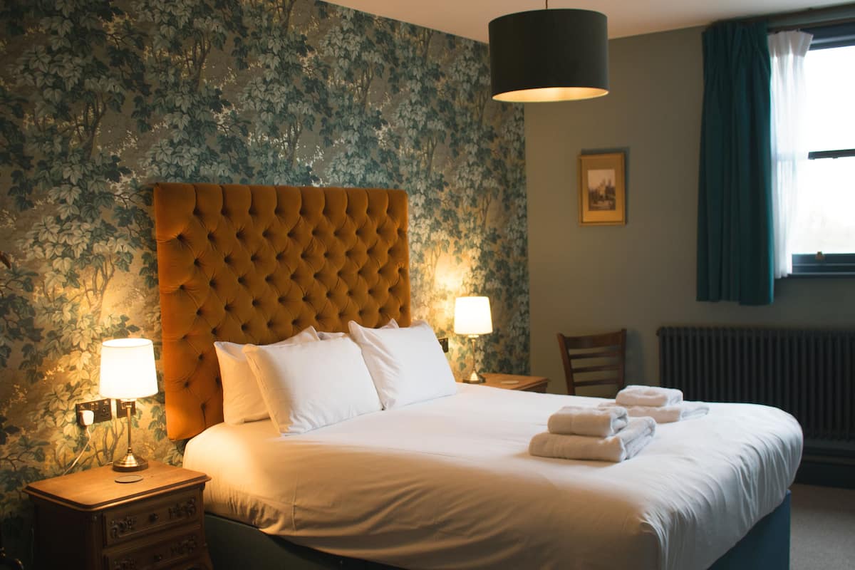 A hotel room with a large bed, two bedside tables, a nightstand, a lamp, a chair, a radiator, and a window with curtains.