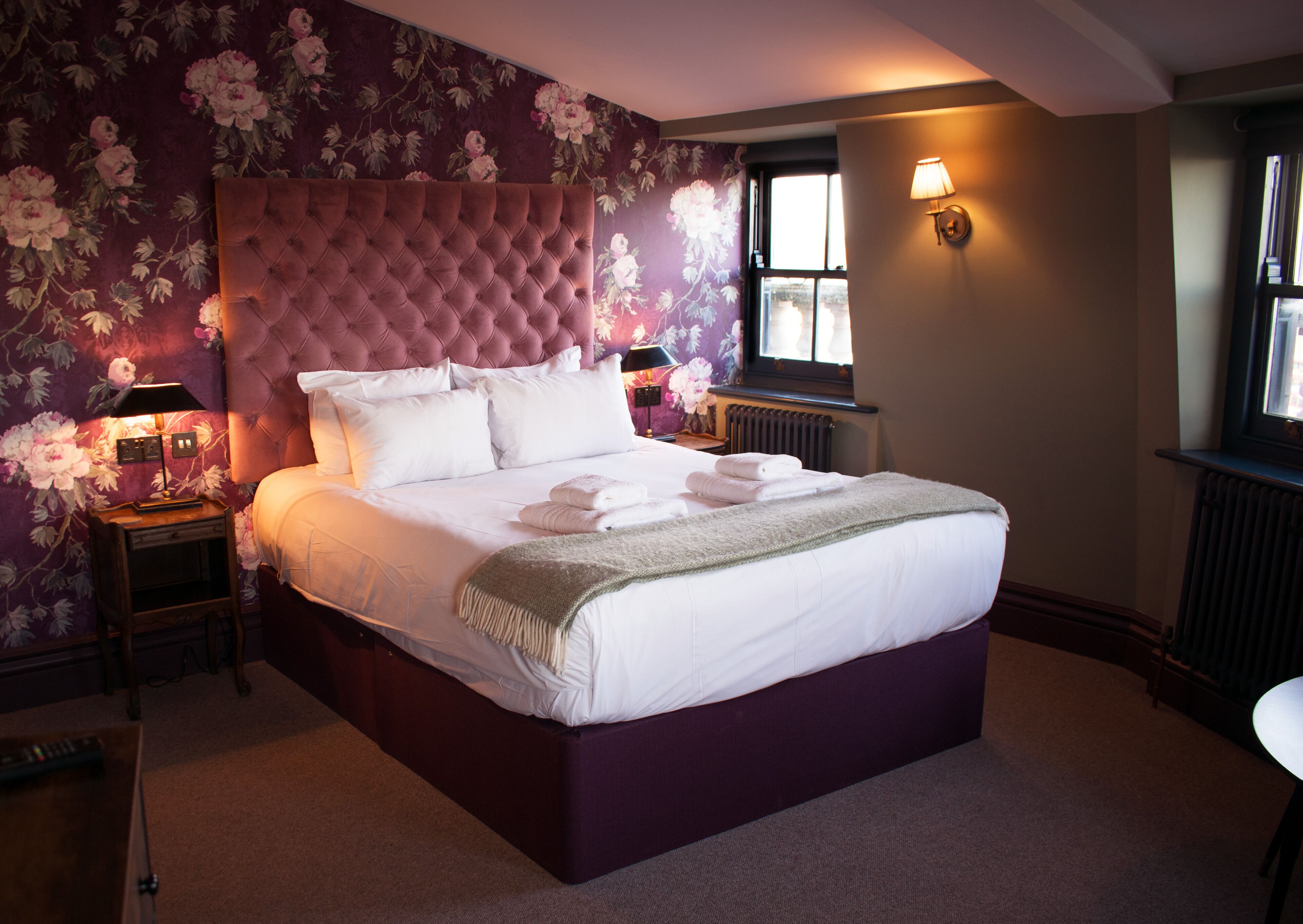 A bedroom with a tufted headboard, a large bed with white linens, two bedside lamps, and a floral wallpaper design.