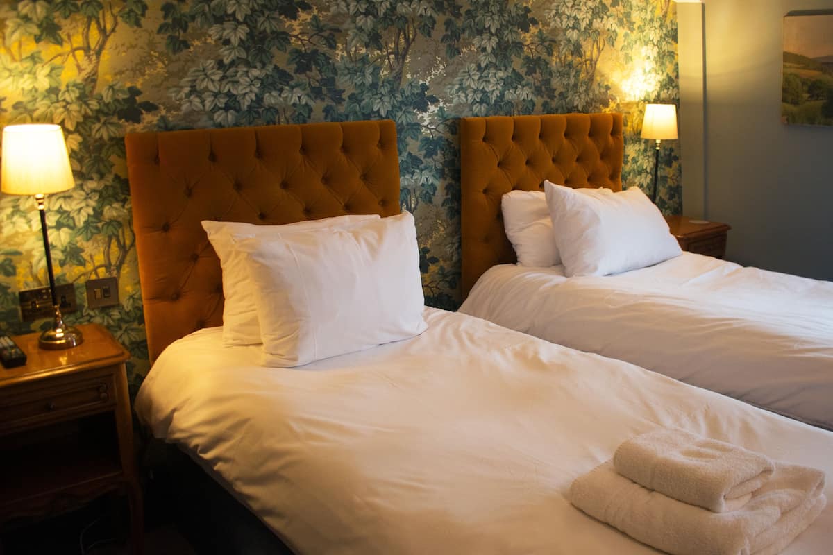 A hotel room with two beds, each with white linens and pillows, and a headboard with a tufted design. There are bedside lamps on wooden nightstands, and a painting on the wall.