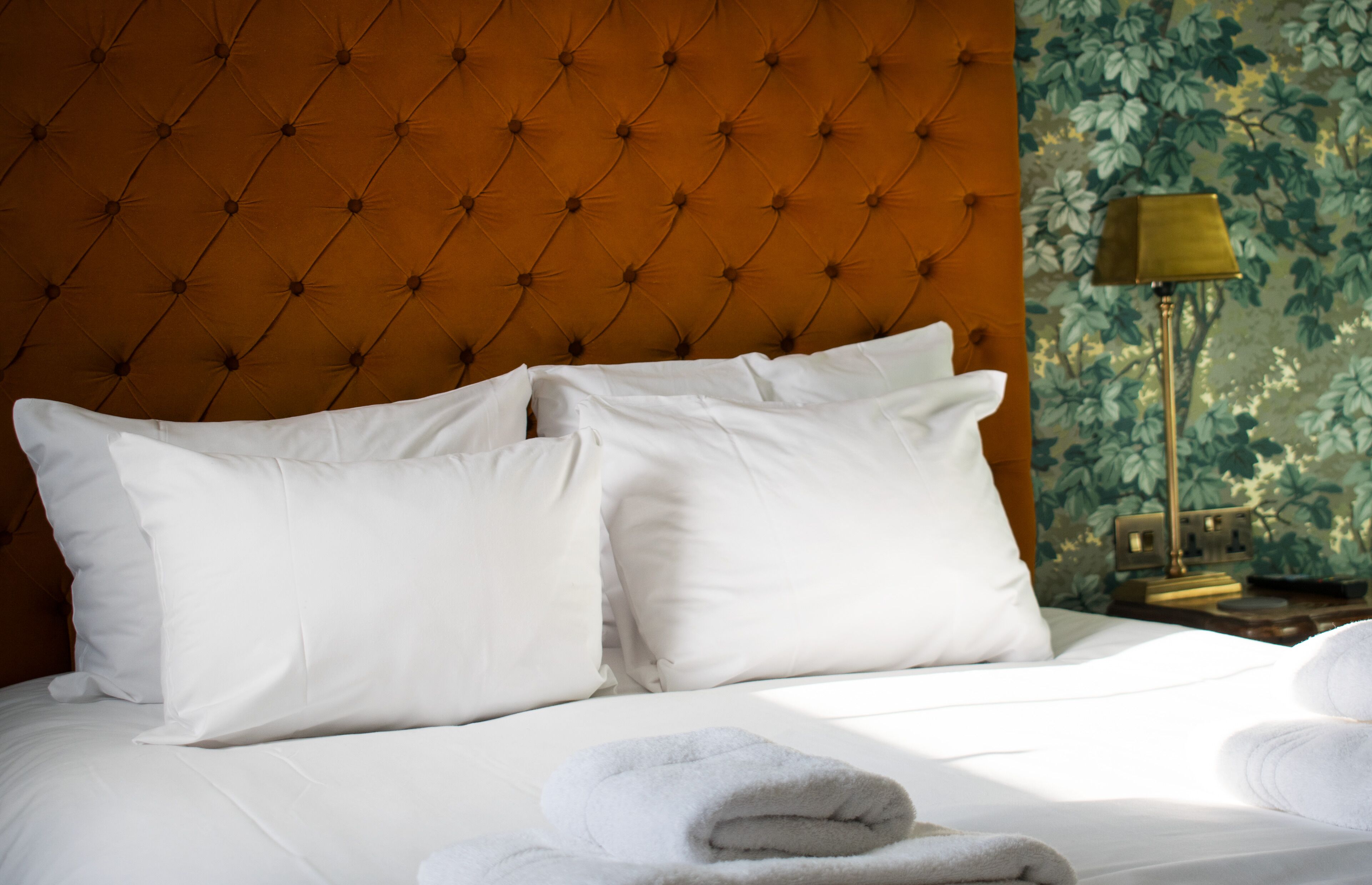 Una cama bien hecha con sábanas y almohadas blancas, cabecera capitonada, una lámpara y papel tapiz con motivos florales.