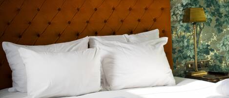 Una cama bien hecha con sábanas y almohadas blancas, cabecera capitonada, una lámpara y papel tapiz con motivos florales.