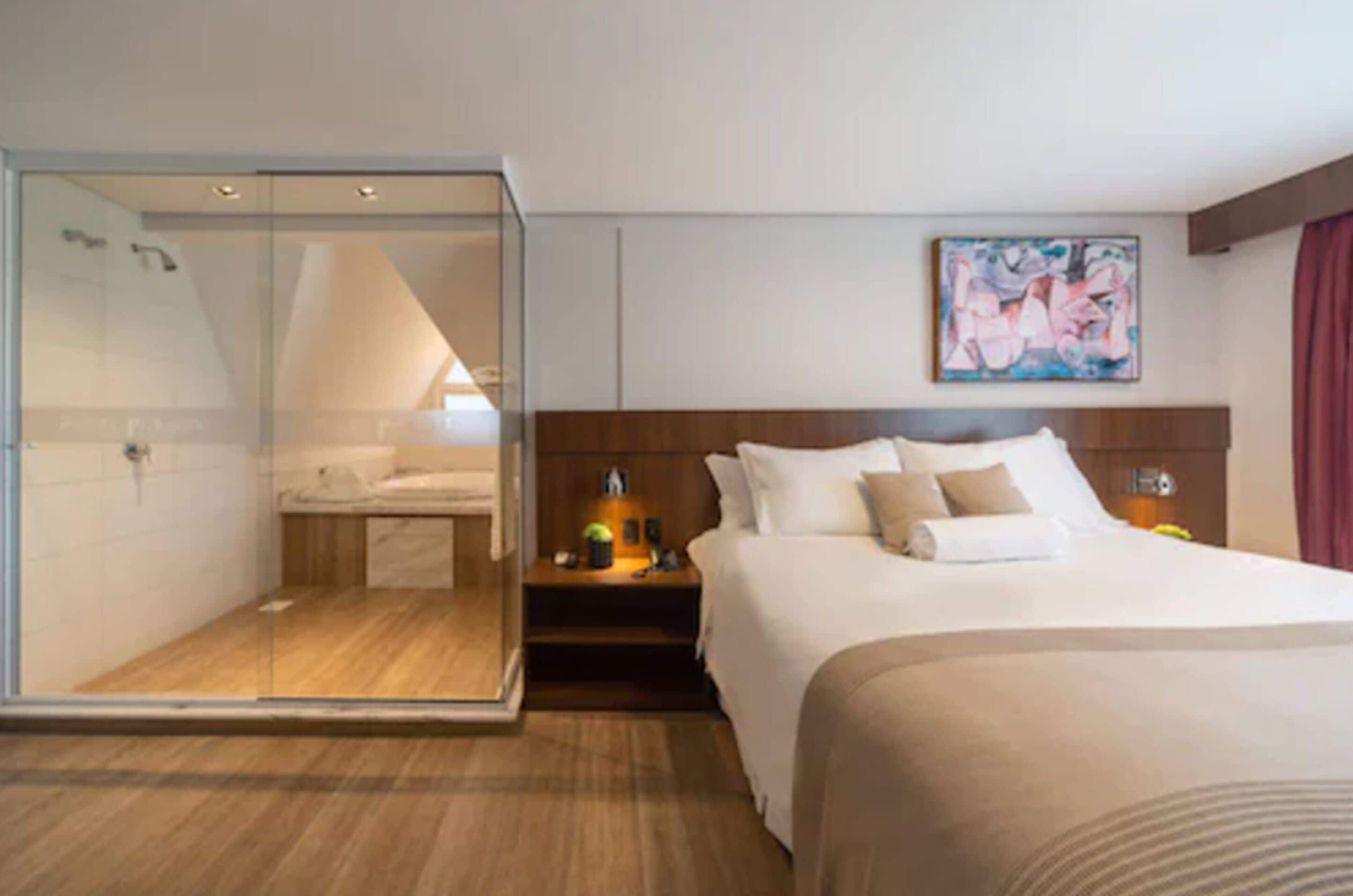 Habitación de hotel con ducha envuelta en vidrio, cama con ropa blanca, cabecero de madera y un cuadro en la pared.