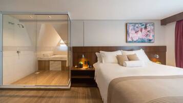 Habitación de hotel con ducha envuelta en vidrio, cama con ropa blanca, cabecero de madera y un cuadro en la pared.