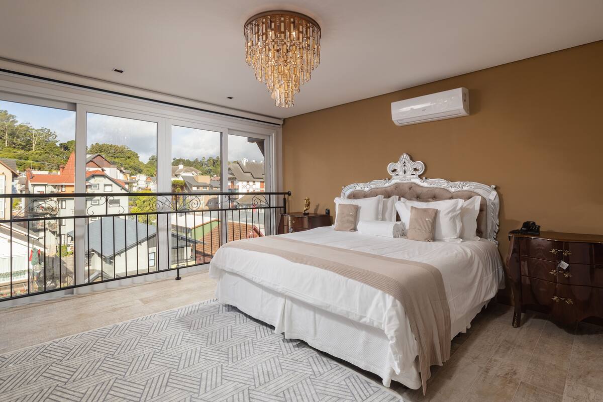 A bedroom with a large bed, a chandelier, a balcony with a railing, and a view of buildings outside.