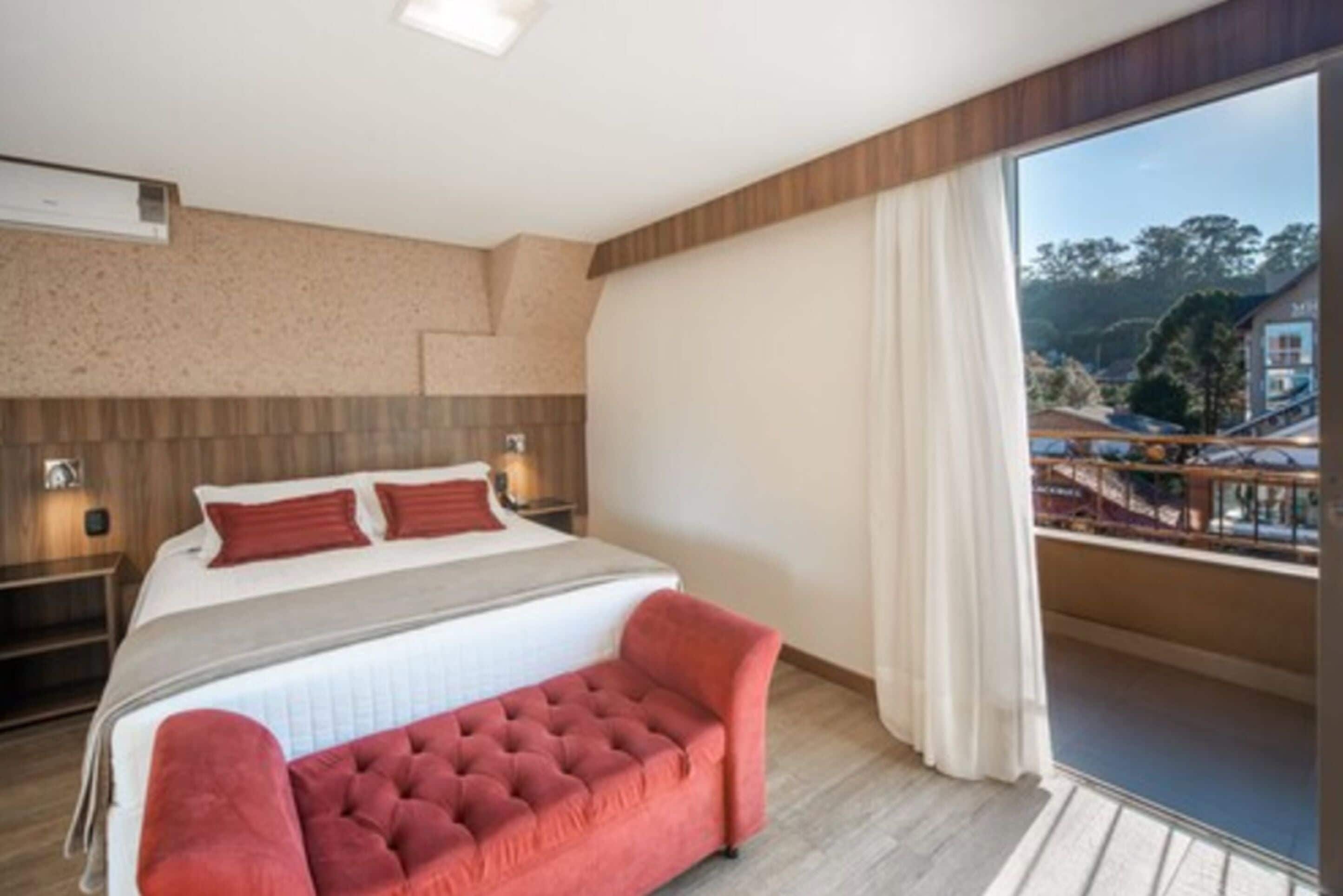 Una habitación de hotel moderna con una cama grande, un banco tapizado de rojo, un balcón con vistas al pueblo y un cabecero de madera.
