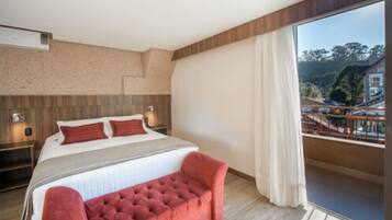 Una habitación de hotel moderna con una cama grande, un banco tapizado de rojo, un balcón con vistas al pueblo y un cabecero de madera.
