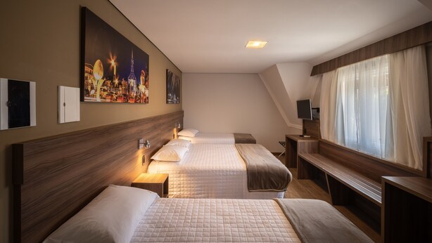 Habitación de hotel con dos camas, un ventanal con cortinas translúcidas, un cuadro en la pared y un escritorio pequeño con una lámpara.