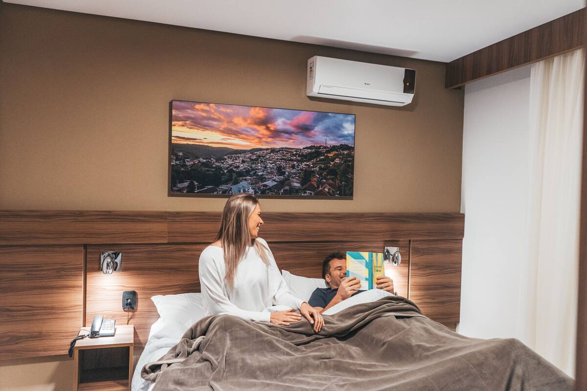 A bedroom with a bed, bedside table with a phone, a wall-mounted air conditioner, and a painting of a sunset over a town.